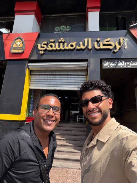 Two friends smile in front of a storefront with Arabic signage and a closed metal shutter.