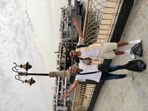 Two travelers pose with arms outstretched on a riverside promenade with Nile cruise ships moored behind.