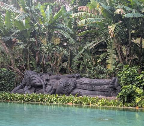 Large reclining stone Hindu deity statue set amid lush tropical foliage in Bali.