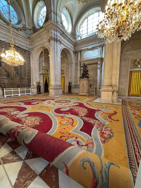 Grand palace hall with ornate yellow and red carpets, marble columns, and chandeliers.