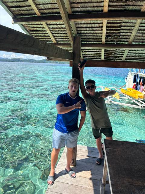 Two friends pose playfully beside crystal-clear turquoise water at an island dock.