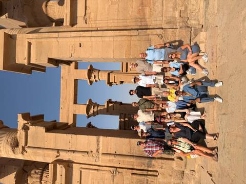 Large tour group posing on the stone steps of an ancient sandstone temple with soaring columns under a clear blue sky.