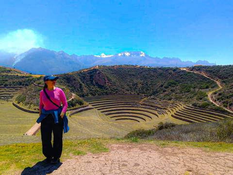 Woman stands before the concentric agricultural terraces of Moray with snow-capped Andes behind.