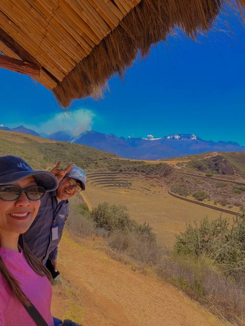 Selfie of two travelers smiling with the circular terraces of Moray and snow-peaked Andes in the background.