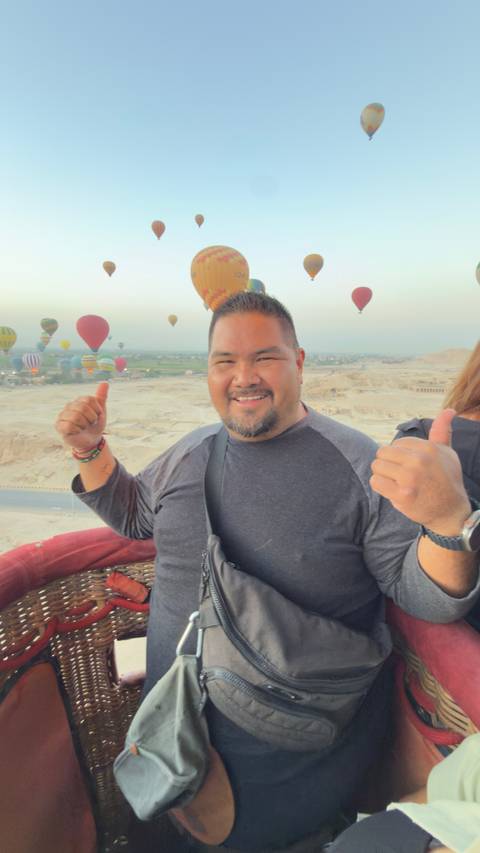 Traveler gives thumbs-up from a hot-air-balloon basket above Luxor with colorful balloons in the sky.