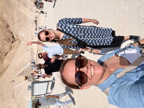 Five smiling travellers pose in bright sunlight on a sandy pathway near ancient stone tombs and cliffs.