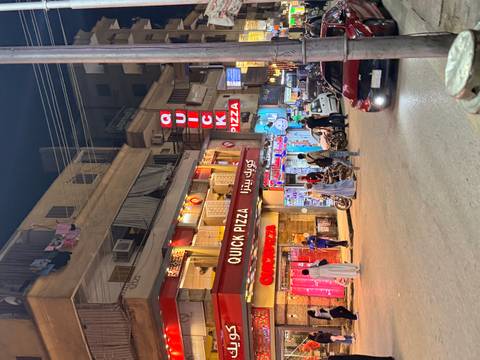 Nighttime street scene with locals and neon shopfronts including a brightly lit 'Quick Pizza' restaurant.