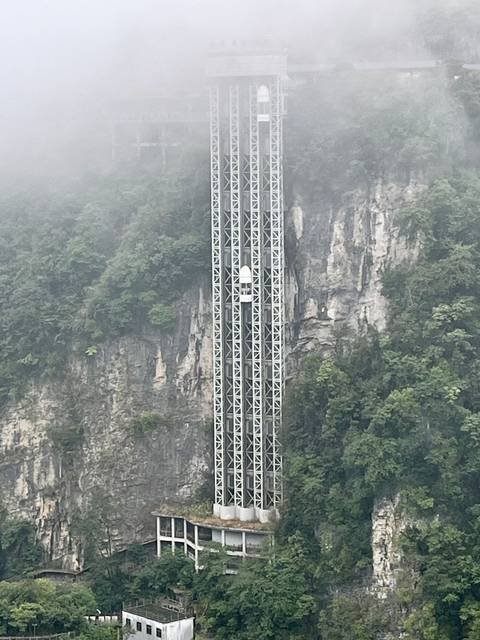 Tall glass-fronted elevator shaft rises against a misty cliff face covered in greenery.