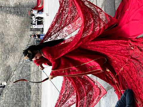 Woman in flowing red traditional costume draws a bow during a performance beside ancient stone walls.