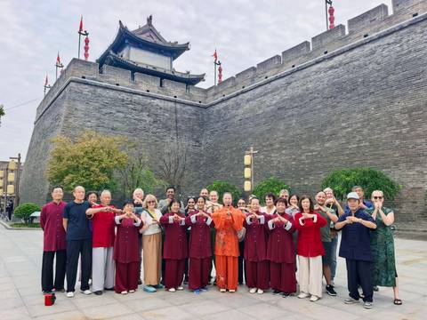 Large group in tai-chi outfits pose with hand gesture in front of a grey brick city wall and tower.