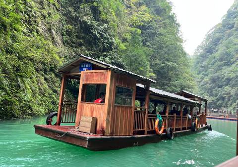 Wooden covered boat with passengers glides along vivid turquoise river between steep forested cliffs.