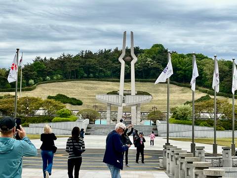 Memorial monument with twin spires and surrounding flags on a terraced hillside, with tourists photographing.