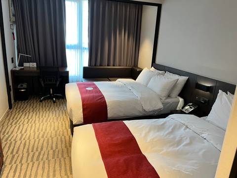 Simple modern twin hotel room with red runners on white beds and work desk by the window.