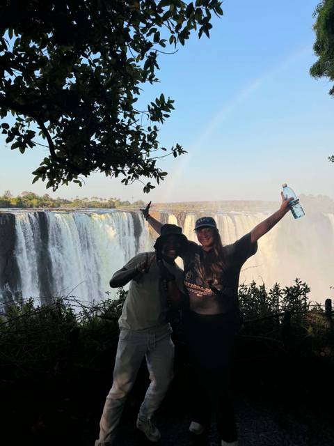 Two travellers celebrate in front of a massive waterfall with mist and a faint rainbow.