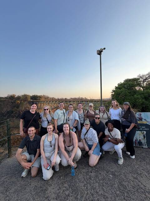 Large tour group smiles at viewpoint with steel railway bridge and canyon in golden evening light.
