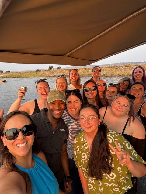 Cheerful boat selfie of travellers with elephants grazing on a riverbank at sunset.