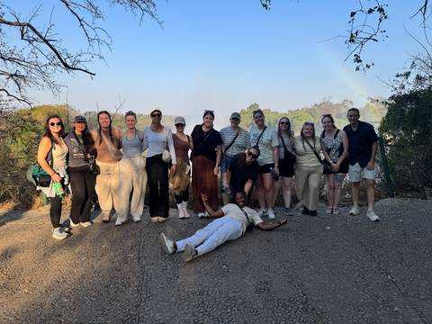 Large tour group posing casually on a dirt path with faint rainbow in the background.