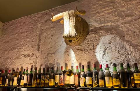 Rustic bar wall with rows of vintage wine bottles beneath a woven straw bull’s head mount.