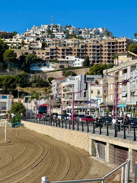 Busy coastal town street with cars, buses and modern apartment blocks climbing a hillside.