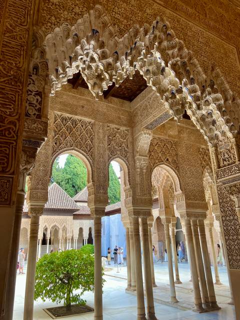 Intricately carved Moorish arches and stucco patterns inside the Alhambra with greenery beyond.
