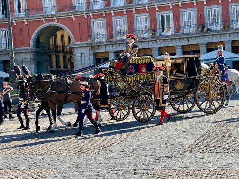 Ornate horse-drawn royal carriage escorted by guards crossing a historic plaza.