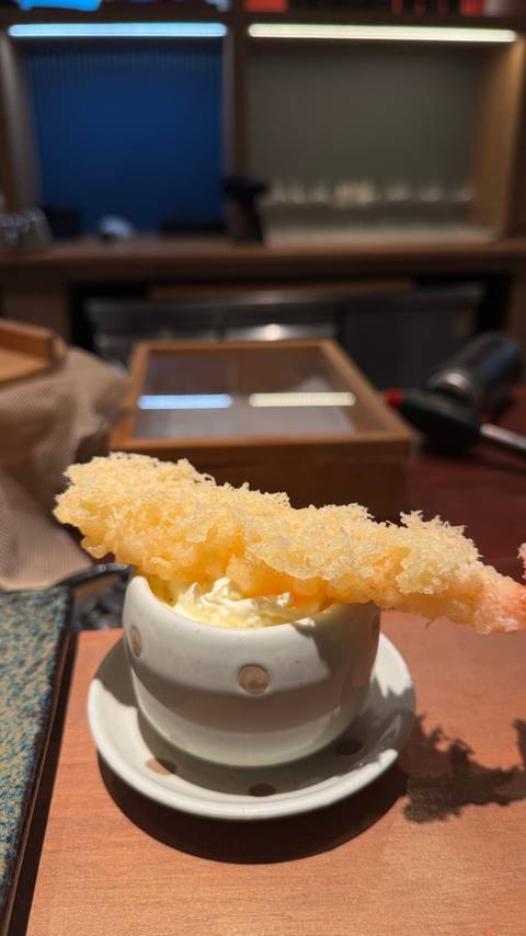 Close-up of a crispy tempura prawn resting on creamy sauce with a blurred kitchen background.