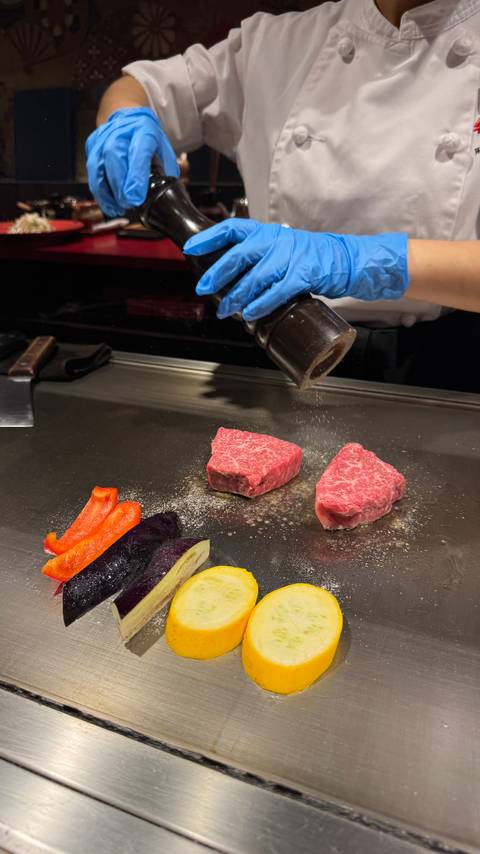Chef seasons raw wagyu steaks on a teppanyaki grill alongside colorful sliced vegetables.