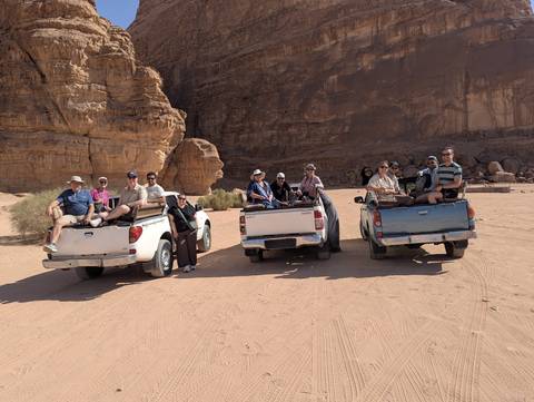 Two pickup trucks with seated tourists parked on Wadi Rum's reddish desert sand beside towering cliffs.