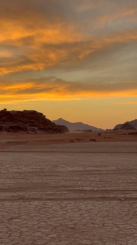 Golden desert landscape and distant jagged mountains illuminated by a vivid sunset sky in Wadi Rum.