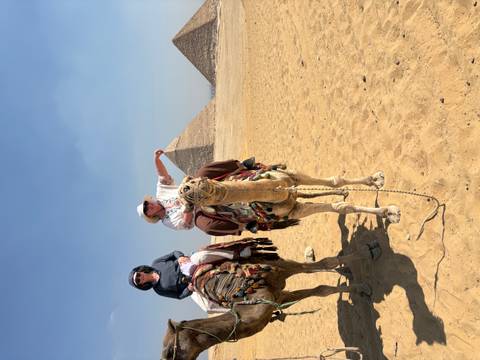 Two tourists riding camels on the sandy plateau with the Giza pyramids rising in the background.