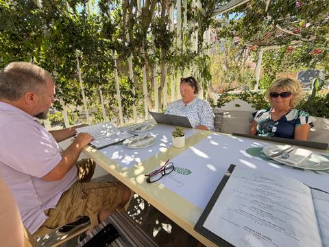 Three travelers reading menus at a shaded outdoor cafe with leafy backdrop.
