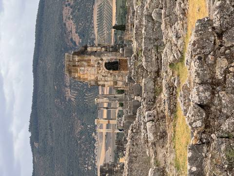 Ancient stone ruins and columns of a Roman archway stand among scattered blocks with green hills behind.