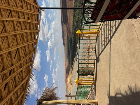 Terrace with bamboo roof frames a sweeping desert canyon and distant palm oasis under a blue sky.