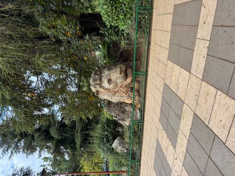 Large carved stone lion head sculpture sits among greenery beside a tiled walkway in a park.