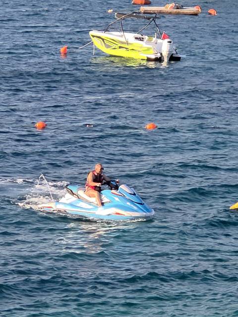 Rider speeds across choppy blue sea on a bright blue jet ski with safety buoys bobbing nearby.