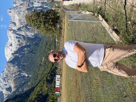 Man stands with folded arms before towering alpine peaks and a white THETH sign on grassy meadow.