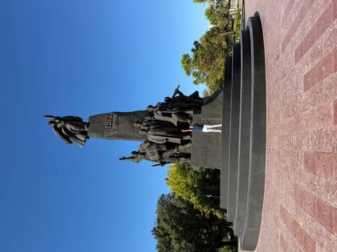Visitor stands on the steps of a monumental bronze statue complex reaching into a clear blue sky.