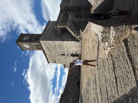 Tourist spreads arms on cobbled ramp before the stone clock tower of Gjirokastra Fortress under patchy clouds.