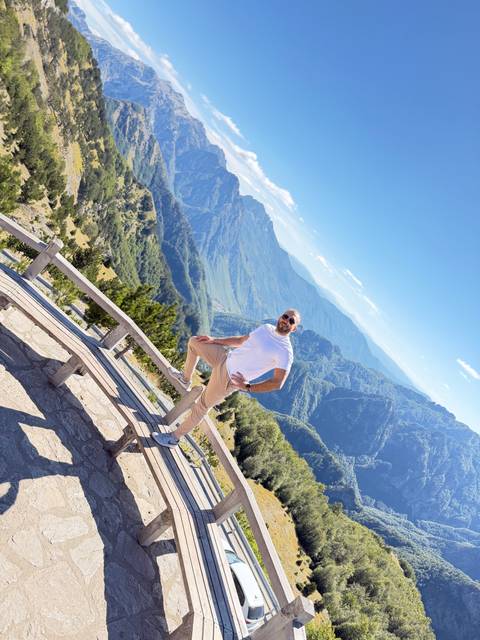Man balances on a wooden bench at a lofty mountain viewpoint with dramatic green valleys below.