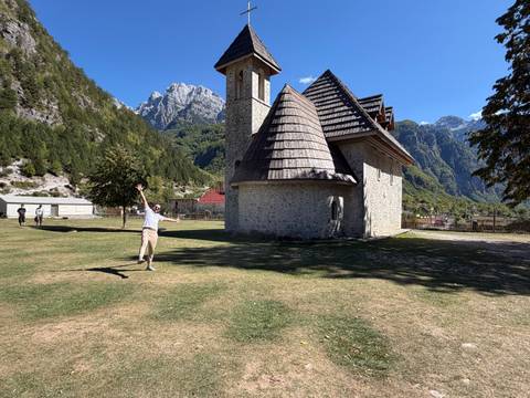Cheerful traveler gestures beside the quaint stone church set amid alpine peaks and blue skies.