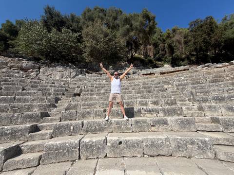 Traveller throws up peace signs while standing in the stone tiers of an ancient amphitheatre surrounded by trees.