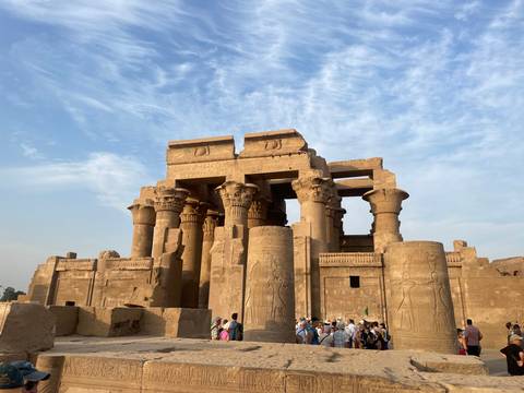 Temple of Kom Ombo with towering sandstone columns rises against a textured blue morning sky, tourists below.