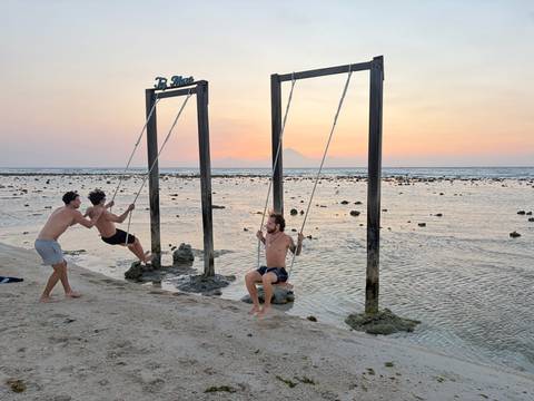 Friends play on iconic ocean swings at low tide with pastel sunset and distant volcanoes.