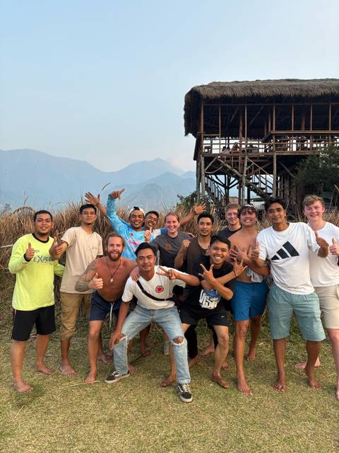 Group of friends smiling and giving thumbs-up on a grassy hillside with hazy mountains behind