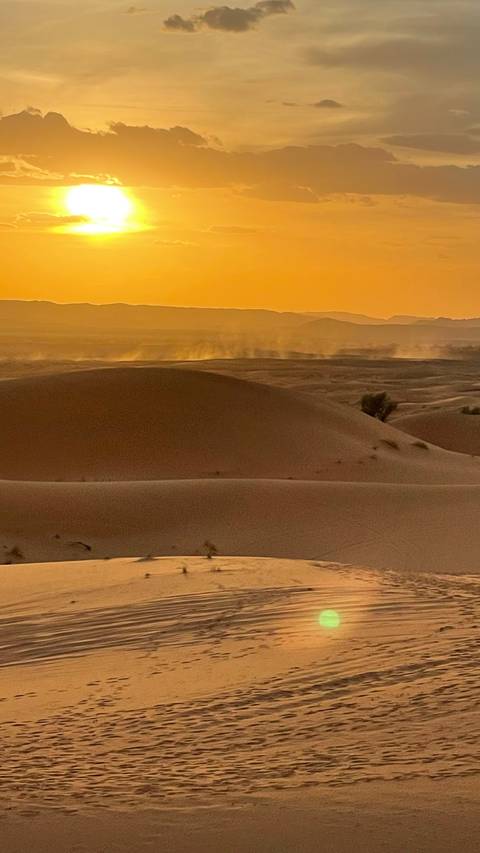Wind-blown sand rises from rolling Sahara dunes under a warm orange sunset horizon.