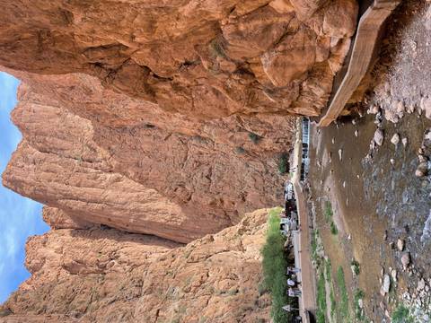 Towering red cliffs of Todra Gorge frame a stream and footpath with vehicles parked below.