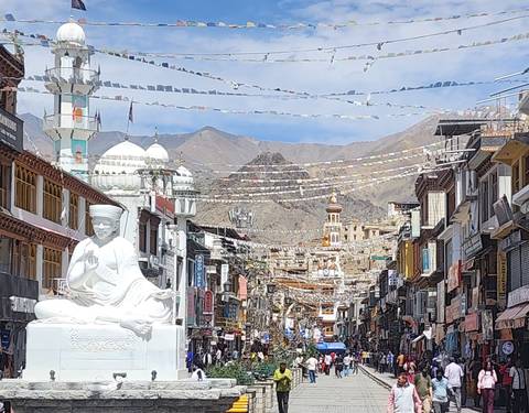 Prayer flags crisscross above Leh’s bustling market street lined with traditional buildings and distant mountains.