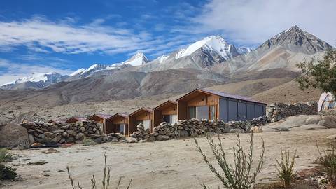 A row of wooden cabins sits in a barren Himalayan landscape below snow-covered peaks and blue sky.