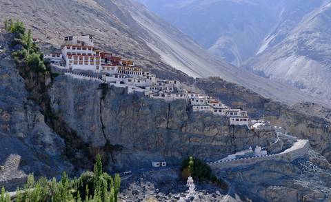 A large white-and-red Buddhist monastery perched dramatically on a sheer rocky cliff with barren Himalayan mountains rising behind it.