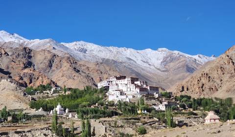 Snow-streaked Himalayan range framing a prominent hilltop monastery and surrounding green oasis villages under a brilliant sky.